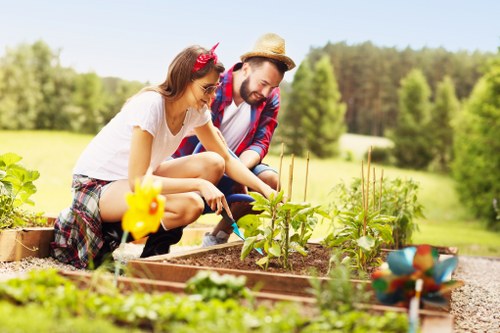 A man and woman are tending to a raised garden bed in a lush, well-maintained outdoor space, which includes a variety of young vegetable plants and flowering plants. The garden bed is bordered by wooden planks, with dark, fertile soil visible where the plants are growing. The woman, wearing a white top and plaid skirt, is using a small gardening tool, while the man, dressed in a denim jacket and straw hat, is engaged in planting or tending to the soil. In the foreground, there are additional garden elements including a colorful ceramic figurine and a decorative planter, while the background features a spacious, green lawn with mature trees and a backdrop of a clear, sunny sky. The scene is bright, indicating good weather, and reflects typical features of a well-kept residential garden in Hammersmith, ideal for outdoor gardening and landscape maintenance services offered by Gardening Services Hammersmith, with a focus on vegetable gardening and outdoor leisure space enhancement.