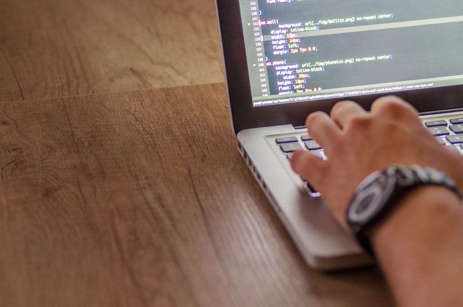A person using a silver laptop on a wooden desk, with their right hand positioned over the keyboard and wearing a wristwatch. The laptop screen displays lines of colorful code on a dark background, in
